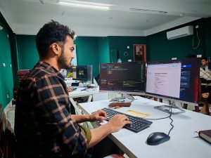 A software developer working on code at a dual monitor setup in a modern office.; VS Code agent sandboxing